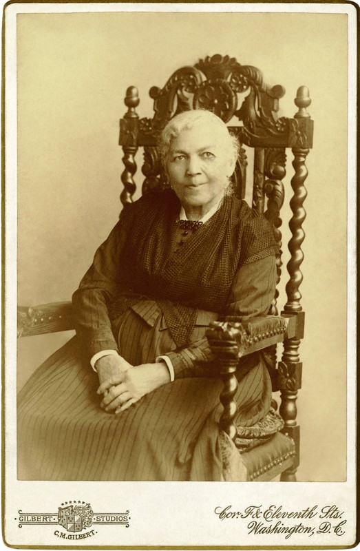 Sepia-toned photo of an old woman sitting in an ornately decorated chair, her hands folded in her lap.