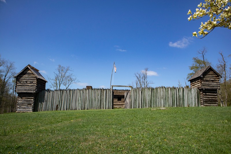 Pricketts Fort is shown directly on, with the doors open that show a log building inside. There are log stockade walls, two out of four corner blockhouses visible. The stockade walls are pointed at the top. The blockhouses are built with logs and clay chinking in between; they have a gabled roof and small lookout windows. The sky is bright blue.