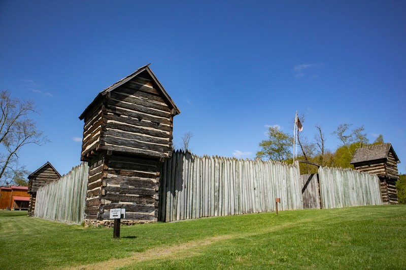 Pricketts Fort is shown from a side view. There are log stockade walls, two out of four corner blockhouses visible. The stockade walls are pointed at the top. The blockhouses are built with logs and clay chinking in between; they have a gabled roof and small lookout windows. The sky is bright blue.