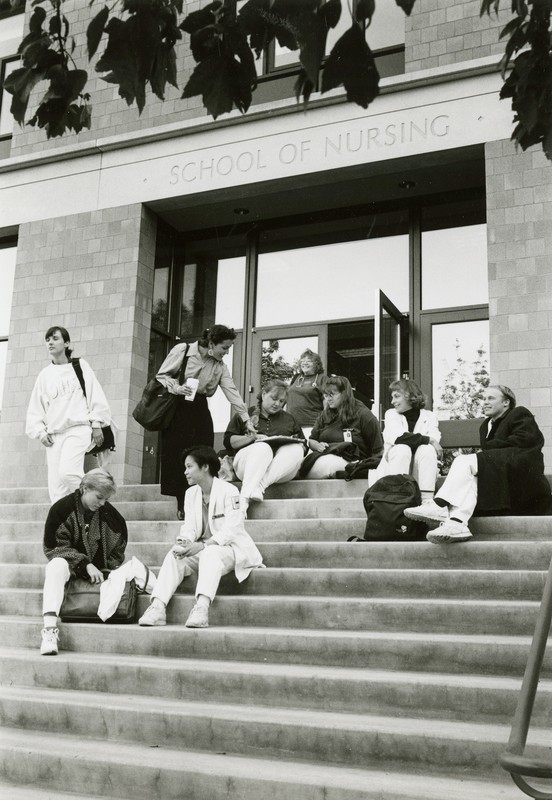 Black and white photograph depicts a group of students sitting on concrete steps leading up to a modern glass entrance in a brick-facade building. The words "School of Nursing" are visible over the doors.