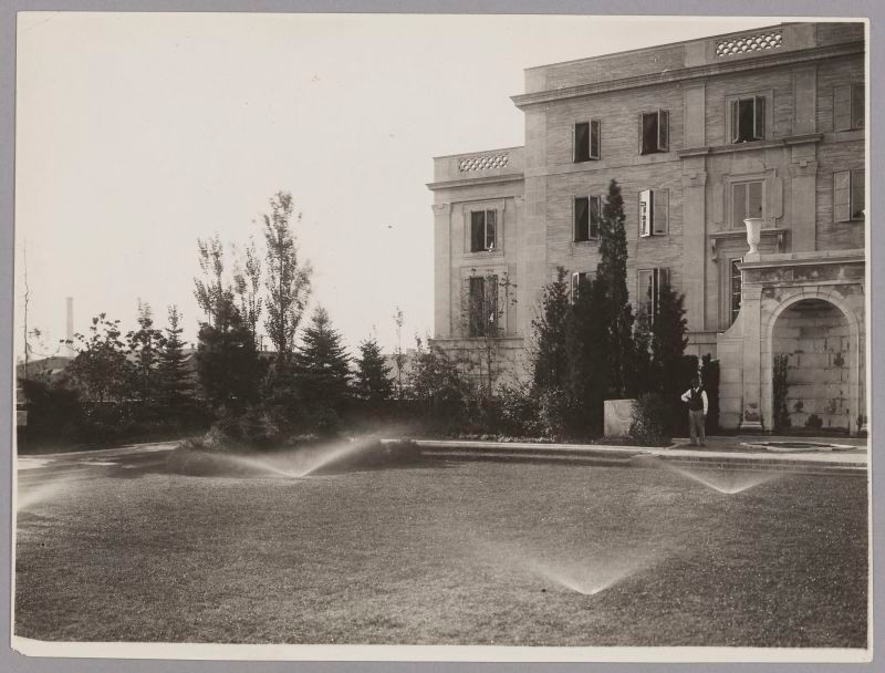 Black & white, building overlooking courtyard garden, Trees border the wall. Water Sprinklers active across the lawn.