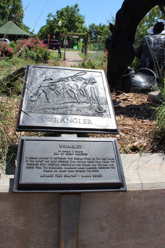 Bronze plaque with a relief image of a man standing beside two horses, throwing a lasso at one of them. Another plaque below it has text explaining the image.