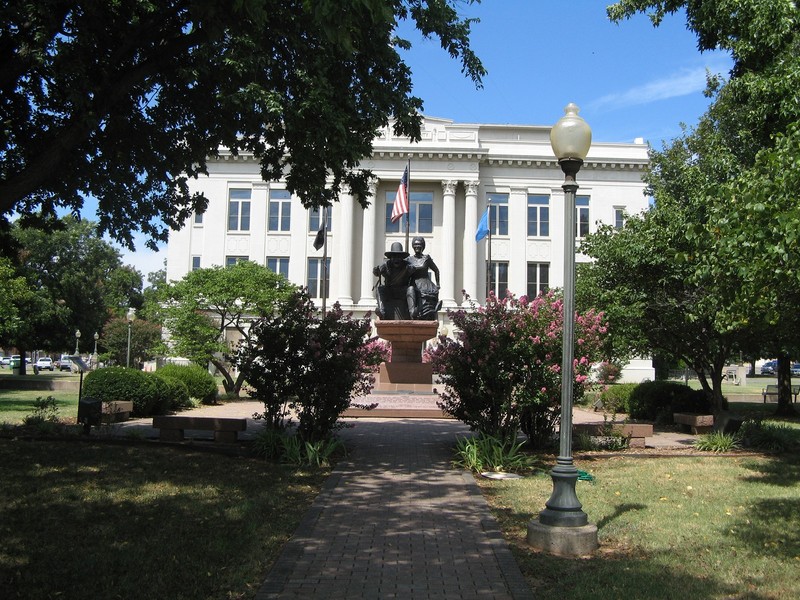 Statue of seated man and woman on stone base in front of large white building with columns on the front.