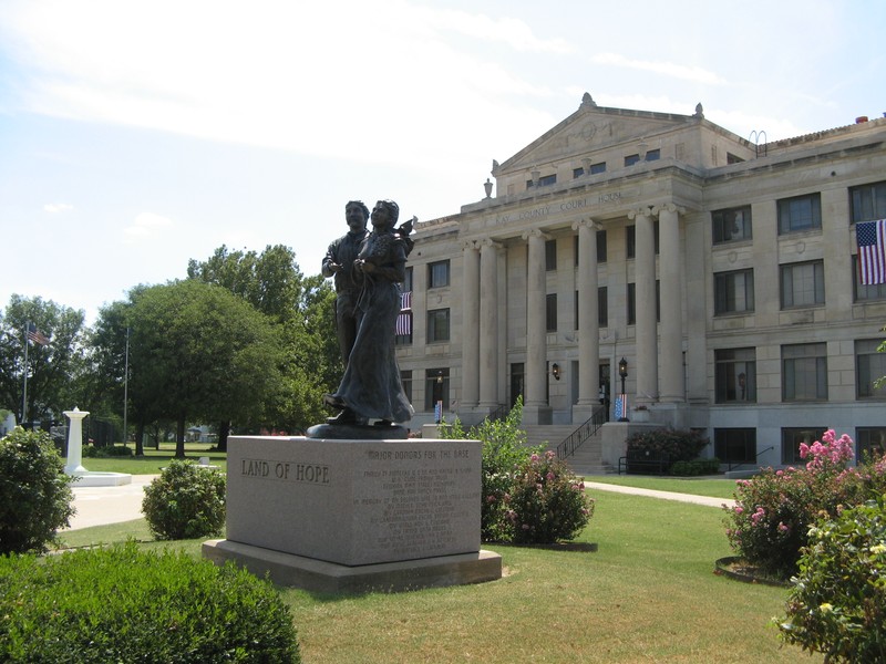 Bronze statue of man and woman on a stone base. Stands in front of a large white building with columns in front.