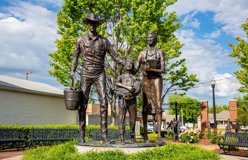 Bronze statue of man in cowboy hat holding a bucket of peaches, woman in buckskin dress holding a book, and young boy holding a rooster