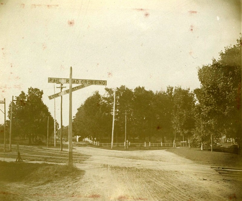Photograph of railroad tracks and crossing.  In the background is Perkins Manor (partially obscured by trees).
