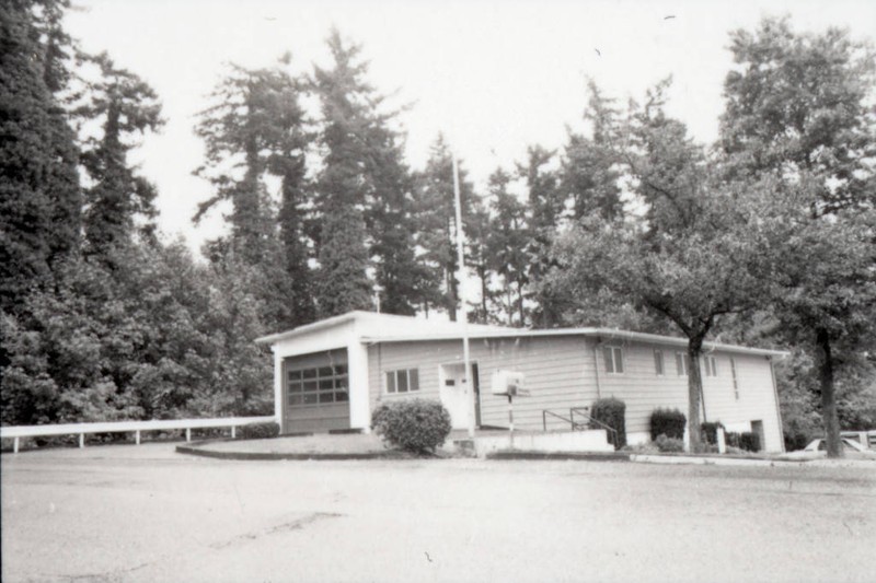 Black and white photograph depicts a one-story building with a large garage at left, at street level with a stand of evergreen trees behind it and a flag pole in front.