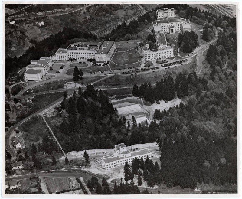 Black and white aerial photograph of the Marquam Hill Campus of the University of Oregon Medical School, Portland, OR in 1940. From left to right: Library, Mackenzie Hall, Doernbecher Memorial Hospital for Children. In the foreground: University Tuberculosis Hospital. Just to the front and left of the Tuberculosis Hospital, the wooden foot bridge crossing the canyon can be seen.