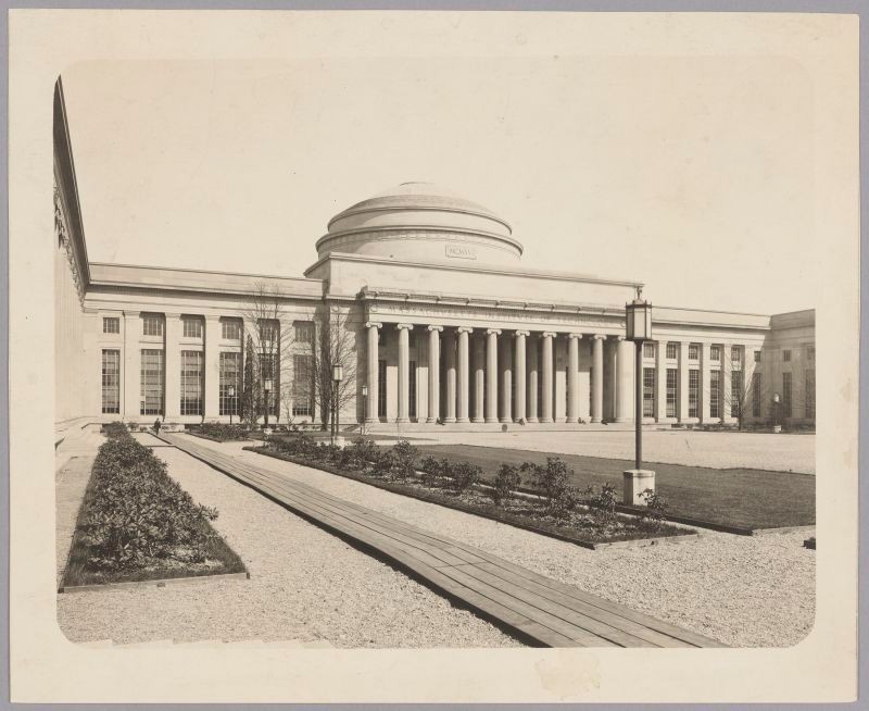 Black & white, domed building with columns across the facade. Gravel ground cover with wooden plank walkways. Planting beds parallel to walkways with young rhododendrons. Leafless trees along the building.