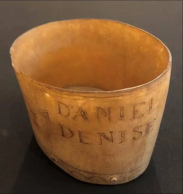 A round cup constructed from cow horn, with two bands carved around the circumference of the top edge and various carvings around the cup such as a two cross-hatched bowtie-shaped designs, a long-tailed bird, and a profile of a bird in flight. "DANIEL / DENISE" is inscribed on the cup's vertical side. There is also a very faint carving of a flower enclosed in a circle. Along the bottom circumference are two bands separated by a scalloped ring. Six iron nails secure a wooden bottom to the body of the cup. On the underside is carved the year, "1779."