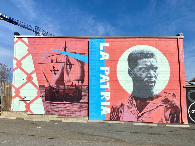 A red, white, and blue mural of a historic tall ship, a young black male figure and the words La Patria.