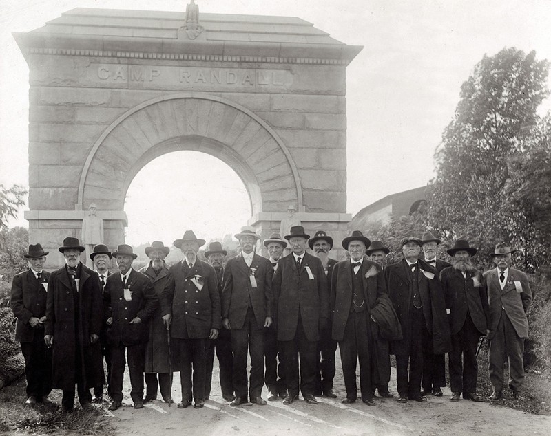 Group portrait photograph of Civil War Veterans standing in front of the Camp Randall Memorial Arch, located in Madison, Wisconsin The veterans are dressed in civilian suits, wearing ribbons and badges, likely from the Grand Army of the Republic. 