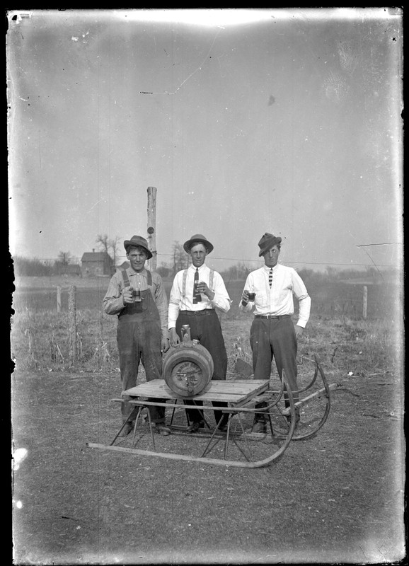 From our collection: three young men, outdoors, each with a mug of beer in their hand. They are behind a sled on which a wooden keg of beer is sitting.