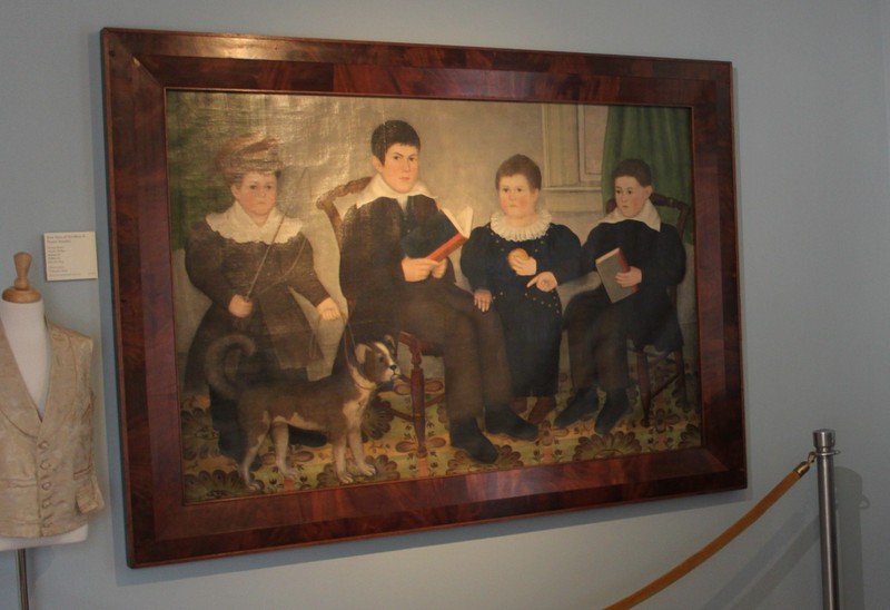 Portrait of Four Boys (American, First Half 19th Century) Unsigned. Two boys are holding books, one is holding an apple and one is holding a riding crop and standing by a dog. The background is a parlor with a patterned rug on the floor. The boys are purportedly George Henry, Charles Wilbor, Stephan H., William E. and Fido the Dog (the name of the dog is painted on the dog in the painting); the boys are the sons of Needham and Naomi Standart.