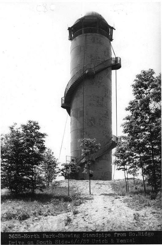 Black and white image shows a tall structure with staircase winding up to a domed roof above an open deck. The deck has scaffolding visible around its outer edge, and ropes are hanging from it to the ground. In the foreground are trees of various ages and size.