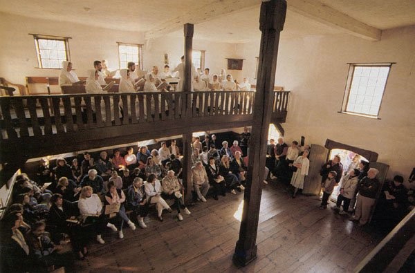 A tour group sits in the house of worship while the choir sings overhead. 