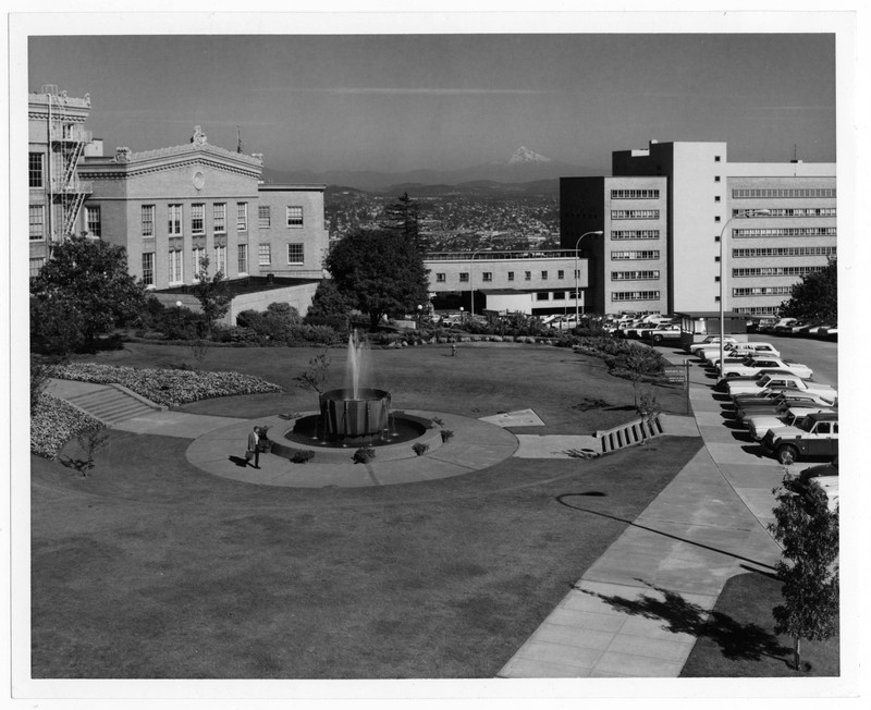 Black and white photograph of Marquam Hill Campus centered on the 75th anniversary fountain in front of Mackenzie Hall and looking east. Part of Mackenzie Hall and Dillehunt Hall visible on the left hand side of the image, with a partial view of University Hospital South (OHSU Hospital) on the right. Mt. Hood visible in background.