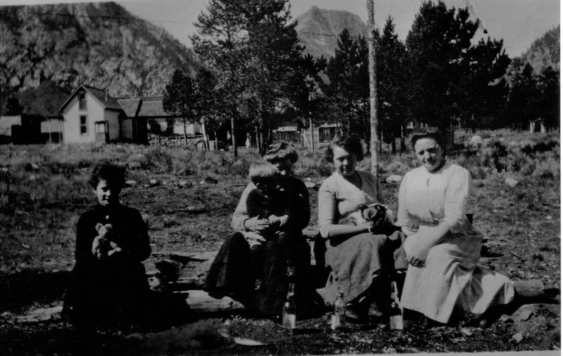 A Black and white photo, circa 1915, featuring a group of women with a young child and a dog. One woman sits apart from the rest holding what looks like a teddy bear. Notice their long skirts and sleeves.