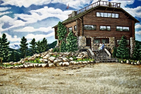 A large, rustic, log cabin sits to the right of the image. Two women sit on the rock stairs in front of the door to the lodge. Several trees surround the house and large rocks form barriers between the grass next to the lodge and the gravel in front. 