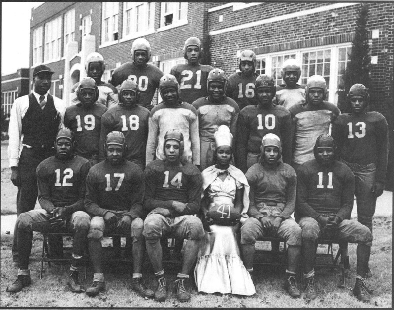 Three rows of football players in uniform along with a homecoming queen in a crown.