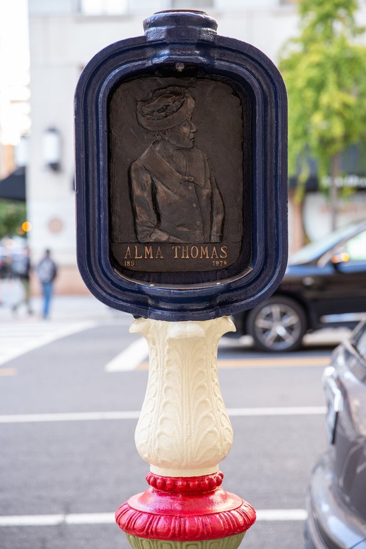 The top half of a sculpture created by repurposing an old emergency call box is pictured in the foreground, a city street in the background. The call box artwork consists of a bronze relief portrait of a woman with the name "Alma Thomas" below it, a dark blue border, and a curvy white and red post.