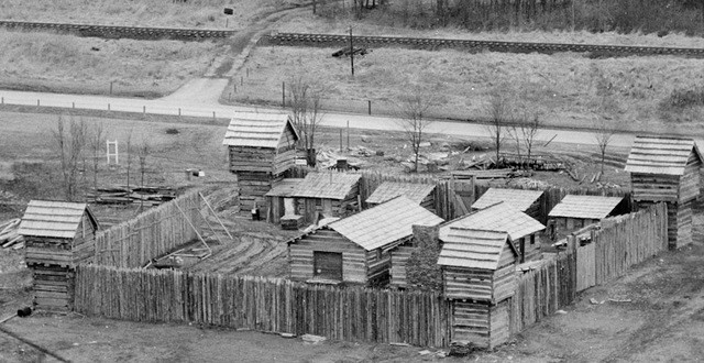 A black and white, aeral image of Pricketts Fort that shows the layout of the buildings inside the fort. The blockhouses are in each corner of the fort and raised high above the stockade walls. The cabins line the interior walls. The larger Trading Post and Meeting House are located in the central green space of the fort.