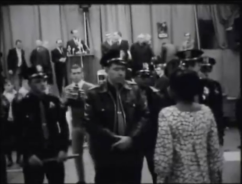 A protestor stands in front of several armed police officers protecting Alabama Governor George Wallace during a speech in Omaha, NE.