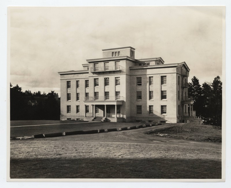 Sepia photograph of an exterior view of the Nurses Dormitory on the Marquam Hill campus, Emma Jones Hall. The building is a four-story classical style building with a short porch on the front. Around the building is a clearing of grass with evergreen trees in the background.