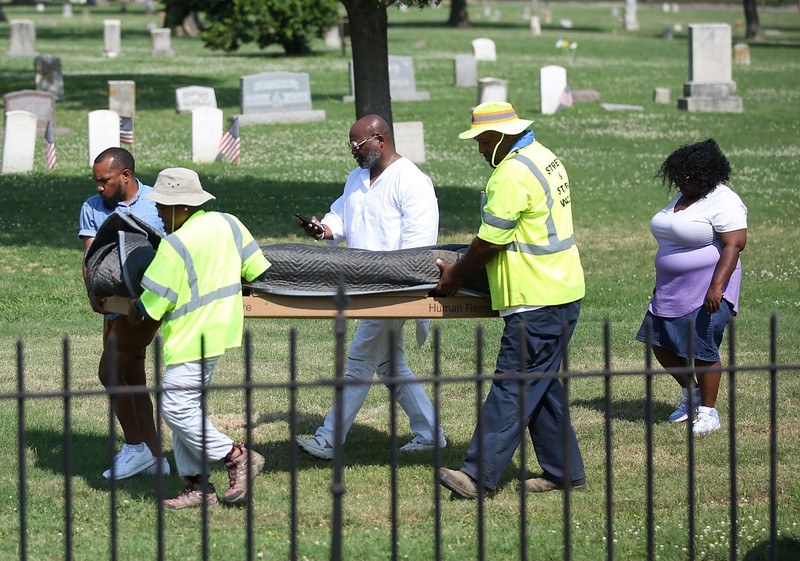 Four people carry a covered coffin or plank in a cemetery
