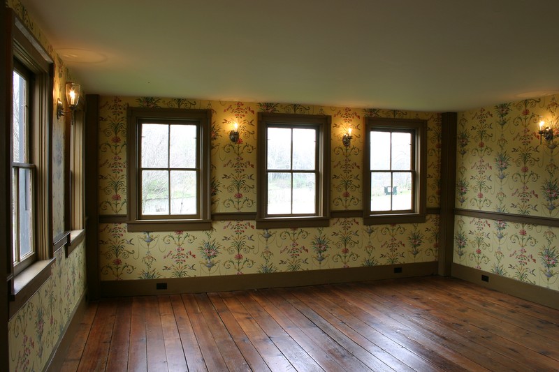 The interior of a room. Yellow floral wallpaper covers the wall, and the floor is bare hardwood. There are three windows. 