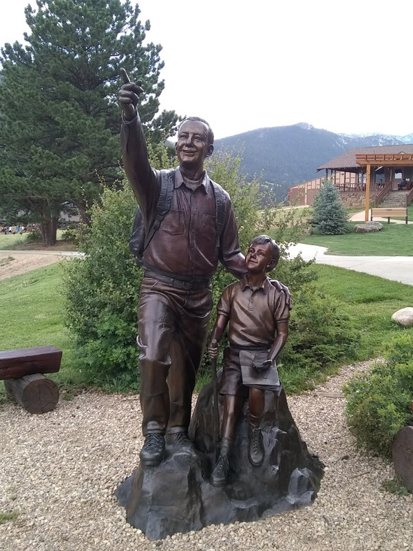 This bronze statue shows two figures. One is a man wearing a backpack, raising his right arm to point at something in the distance. The second figure is a young boy sitting on a rock next to the man with a map and compass on his lap. The boy watches the man as though they are having a conversation.