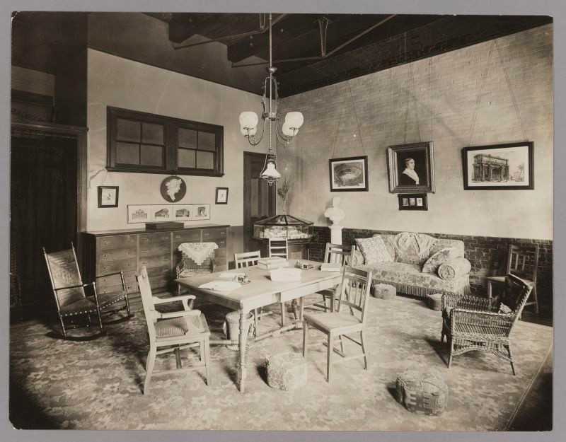 Black & White photo. Room with chairs around a central table, chairs and upholstered sofa. Dark wainscotting.