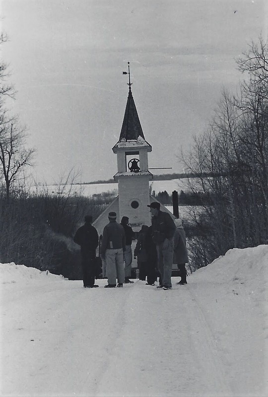 Men in winter clothing observing as the church is moved along a steep hill on a snowy road.