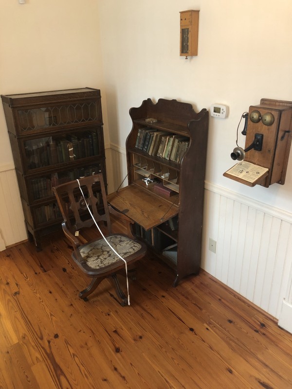 This is Dr. Pope’s home patient examination area. Moving from left to right there is a bookcase that contains Dr. Pope’s original medical books (pre-penicillin). Dr. Pope’s original desk and chair, hanging above it is a maid’s call box, and to the right is a replica of the model of phone the family would’ve had.