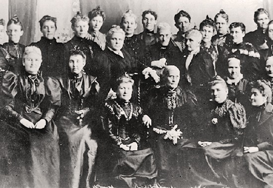 Susan B. Anthony, seated in center, wearing spectacles, and Anna Howard Shaw, standing at the left with her hand on a chair, met with a group of Utah woman suffrage leaders in 1895 after they united both of their organizations..