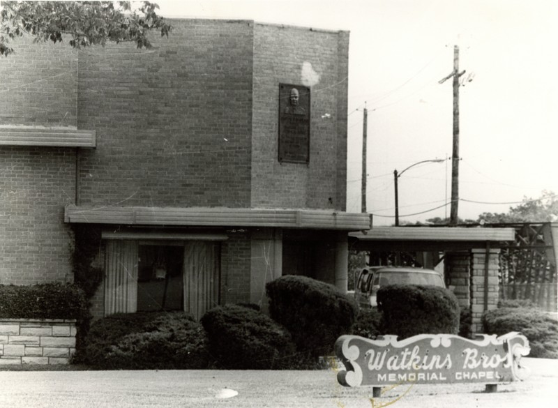 The image shows a two-story brick building. In the center of the image, the doorway of the building is partially visible, though it is obscured behind some hedges. The doorway is situated on the corner of the building. Above the door, there is a decorative elements running around the building. Above the decoration, there is a large plaque on the wall. The plaque has a portrait of a man in the top third, with the rest being text. The text is not legible in this image. On the left side , a window with partially-drawn curtains is visible on the first floor. On the right side, an awning supported by two pillars can be seen extending out from the building to cover part of a circle drive. A van is parked under the awning. In the background on the right side, several power line poles or light posts are visible, as well as an indistinct metal structure on the other side of the street. In the foreground in front of the building and the hedges, there is an open grassy lawn with a sign in it. The sign is medium-color with light-colored scrolling designs around the top and sides and light-colored text. A large line of cursive text on the sign reads "WATKINS BROS" with a smaller line below reading "MEMORIAL CHAPEL".