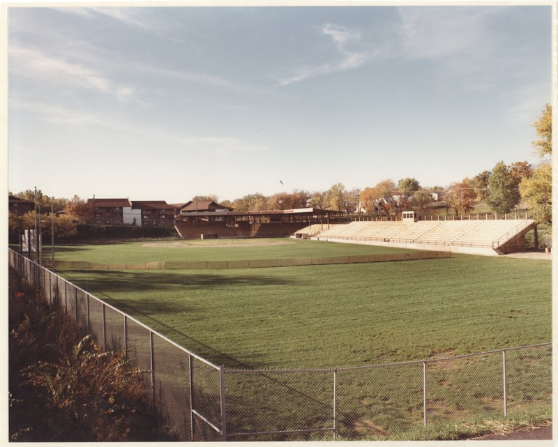 A view of a baseball stadium from the outfield. In the foreground, a chain link fence can be seen, with a large grassy field beyond it. In the background, concrete bleachers with an awning above them can be seen bordering the infield. Beyond the bleachers, trees and other buildings can be seen in the distance. 