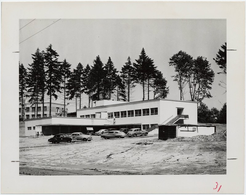 Black and white photograph of the Child Development and Rehabilitation Center during construction. View is of the exterior and parking, set in front of stand of evergreen trees.