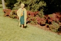 A woman (Alma Thomas) in a green dress, yellow scarf, coat, and hat, and carrying a black bag walks through a grassy area with trees and azalea bushes in the background