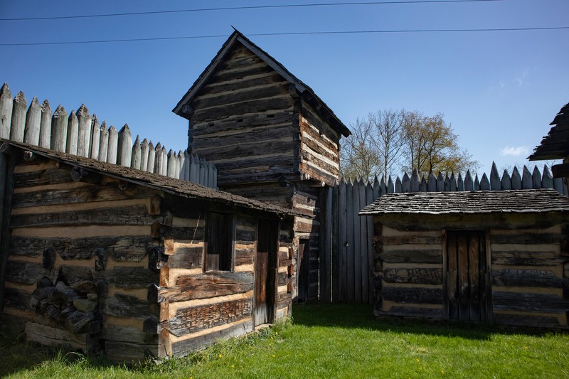 Three small, one-room cabins are visible in this image, along with one of the blockhouses. The cabins have a window or two and open door. The sky is bright blue.