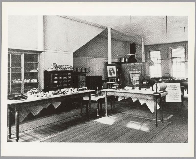 Long wooden tables placed at right angles covered with plates of food. Dishes and pots on cabinets along the wall. Portrait of a man, Count Rumford, in the background.