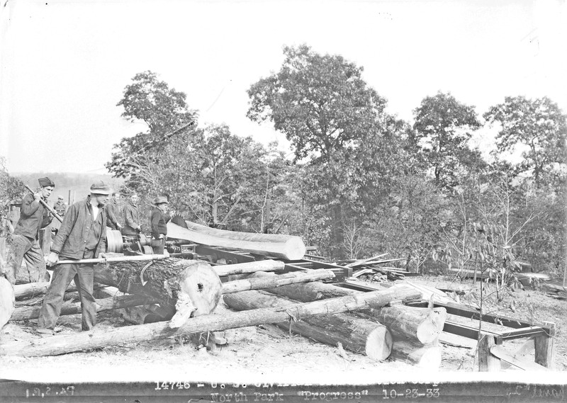 Black and white photo shows six men at work milling logs. Two men use cant hooks to hold a large log on a timber ramp. In front of them one man stands next to a very large saw blade that is slicing a long piece off of another log. The other three men stand behind the saw blade.