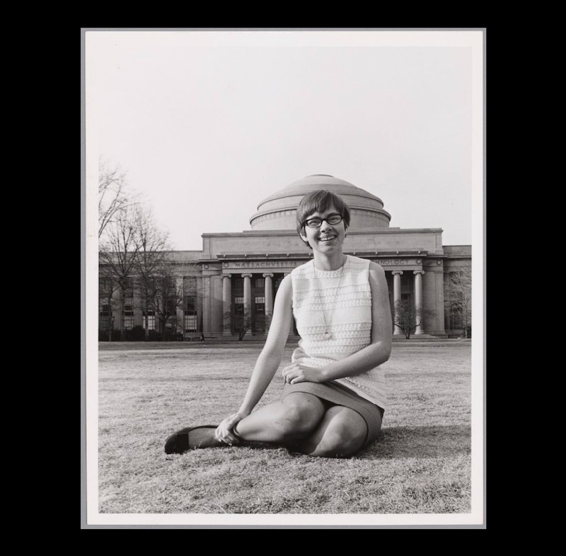 Black & White photo. Young woman with short hair wearing a sleeveless light blouse, and skirt. Sitting on the grass in front of white domed building with columns across the facade.