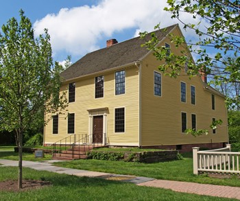 A side view of the Silas Deane House. The two-story home features yellow siding, a brown door, and slanted roof. A small brick stoop leads from the sidewalk to the front door. 