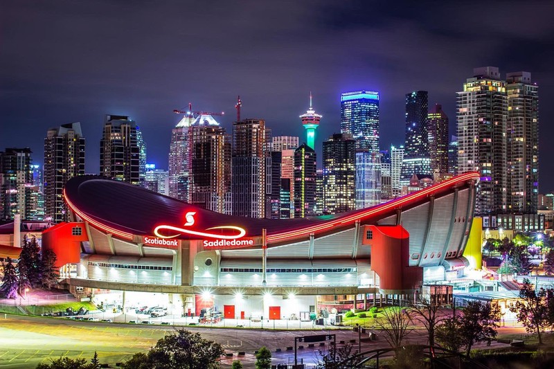 We can see in the centre of the image the building from the front view where you can see the Scotiabank Saddledome in Alberta, Calgary behind it there’s the light-up city of Calgary filled with a skyline of many skyscrapers. The Saddledome was named for its horse-saddle-shaped roof. The arena has a round shape with sort of huge pillars of red and yellow colors on its side. In addition, since this picture was taken at night the building are fully lit up, at the top we can see a band of red light that surrounds the roof of the infrastructure, and then in the front, the Saddledome logo which is a red and yellow infinity sign with the Scotiabank logo above. Then below it around the building's base is covered in white/blue-ish lights. Despite there being 10+ skyscrapers in the background of this photo the way that the Saddledome is lit up and positioned in this photo it appears to be the largest structure standing out more than the buildings behind it. The way this photograph was taken really makes the Saddledome stand out compared to the background, mainly because the Saddledome stands out with red lighting and the background is mostly blue but also because the buildings closest to the Saddledome don't compare in size.