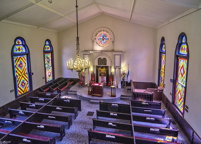 Mahagony pews with red cushions surrond the Bimah, a raised platform, at the front of the synagogue. The Bimah is backed by the flag of Israel and a stained glass window.