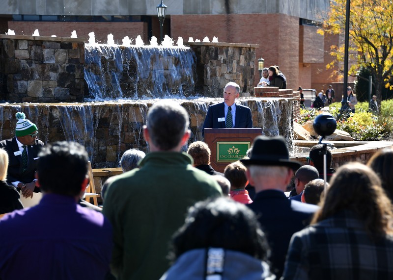 Chancellor Phi Dubois speaks at the Belk Plaza dedication at UNC Charlotte in 2018. The Belk Plaza fountain is behind Dubois and a crowd of people is in the foreground.