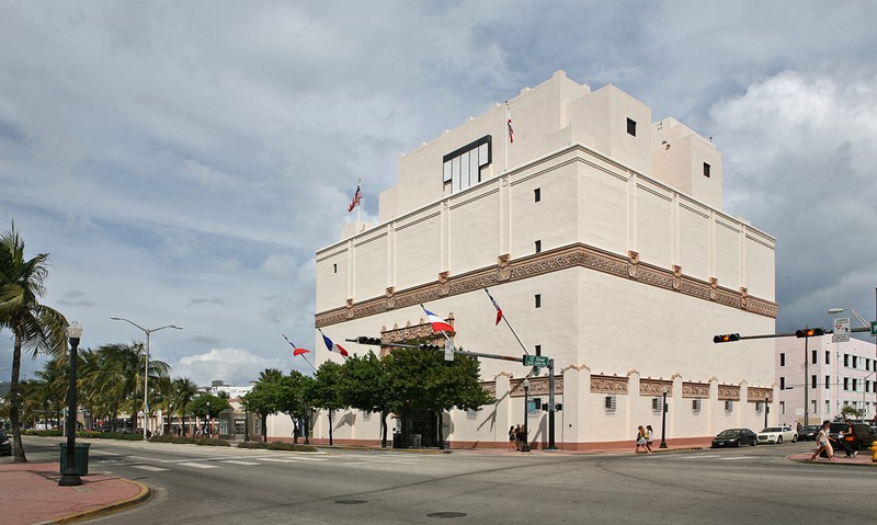 A large creme colored building stands next to a street lined with trees.