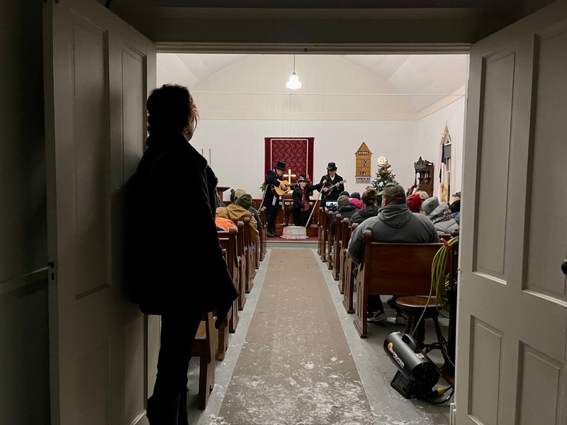 View of the Ebenezer Church interior from the front door. Three men in black suits and top hats decorated with holiday garland are playing traditional instruments. Visitors in contemporary winter clothes are sitting in the pews. The entranceway is dark, with a dark figure seen in silhouette to the left. The interior is well-lit from overhead lighting and an illuminated cross behind the band.