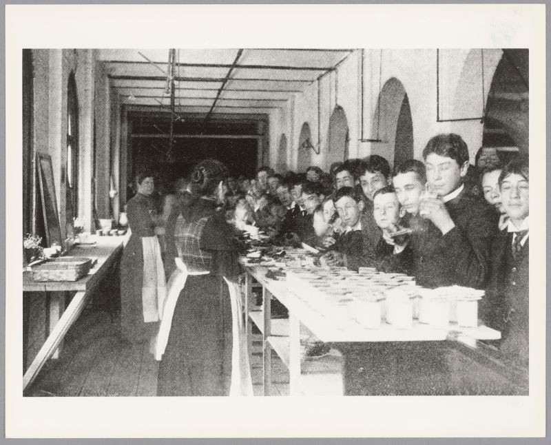 Teen boys gather at a lunch counter at the Boston Latin School. Two women wearing aprons serve food. 